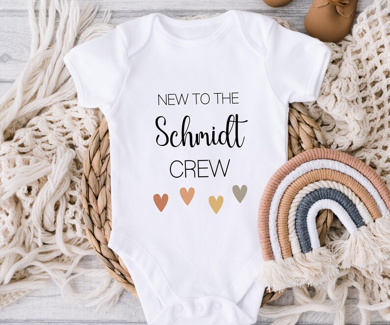 May include: White baby onesie with the text "New to the Schmidt Crew" in black. Five small hearts in different shades of brown, orange, and gray are below the text.
