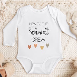 May include: White long-sleeved baby onesie with the text "New to the Schmidt Crew" and five small hearts in shades of orange, brown, and beige.