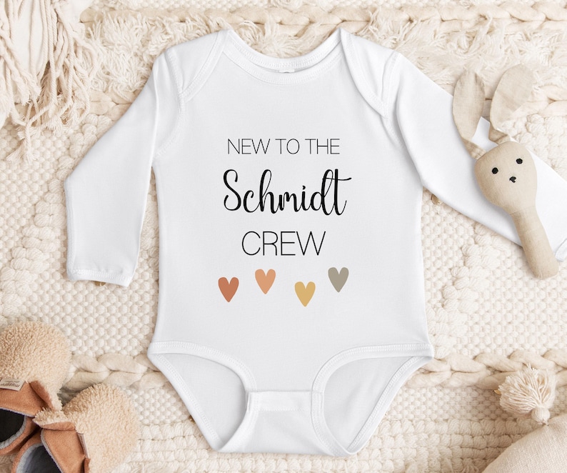 May include: White long-sleeved baby onesie with the text "New to the Schmidt Crew" and five small hearts in shades of orange, brown, and beige.