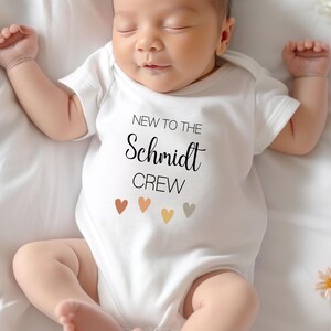 May include: A white baby onesie with the text "New to the Schmidt Crew" and five small, colorful hearts.