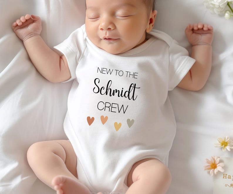 May include: A white baby onesie with the text "New to the Schmidt Crew" and five small, colorful hearts.