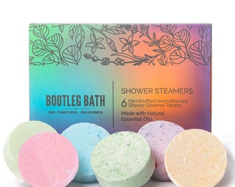 May include: A box of Bootleg Bath shower steamers with six colorful tablets. The box has a gradient background and the text "Bootleg Bath" and "Shower Steamers". The tablets are pastel green, pink, blue, and yellow.