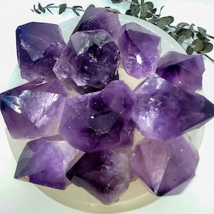 May include: A collection of raw amethyst crystals in varying sizes and shapes. The crystals display a deep purple hue with natural facets, resting on a white, shallow dish. The arrangement showcases the crystals' natural beauty and texture.