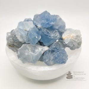 May include: A collection of rough, light blue celestite crystals fills a white, shallow bowl. The crystals vary in shades of blue and gray, with a crystalline texture. The bowl is smooth and white, contrasting with the textured crystals. The image is well-lit against a white background.