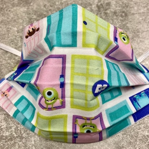 May include: A white face mask with a colorful pattern of doors and characters from the movie Monsters Inc. The doors are in shades of green, blue, and pink, and the characters are Sully and Boo. The mask has elastic ear loops.