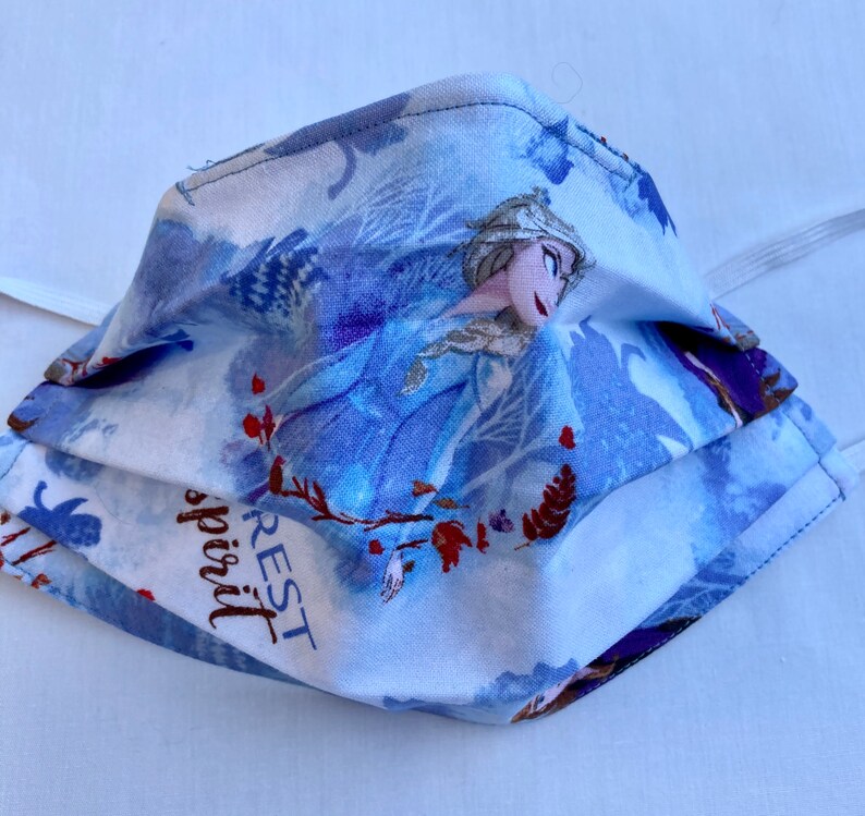 May include: A blue and white face mask with a print of Elsa from the movie Frozen. The mask has the words "Rest spirit" printed on it.