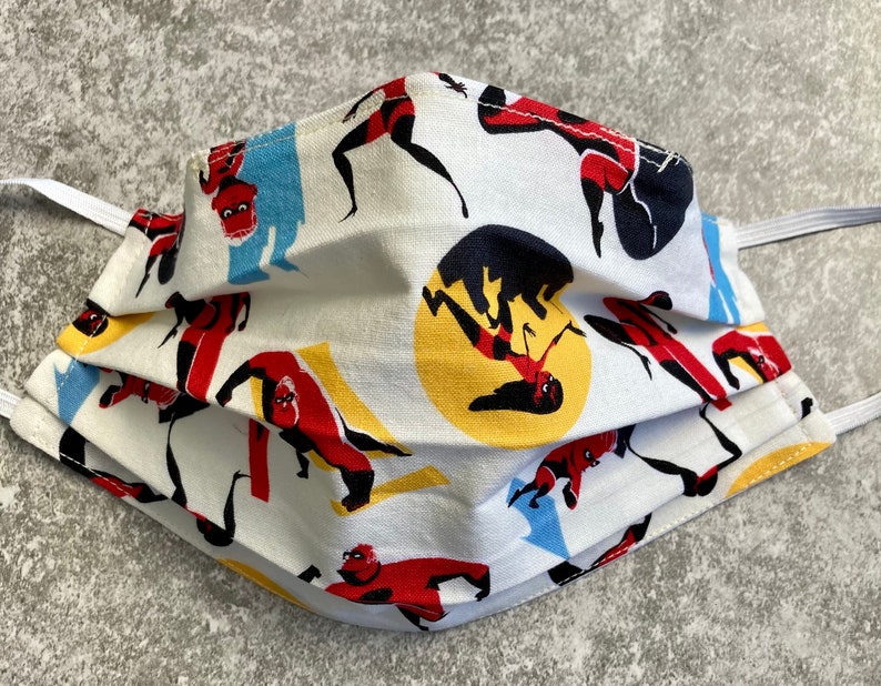 May include: A white face mask with a repeating pattern of a superhero in red, yellow, and black. The superhero is in various poses, including flying and punching.