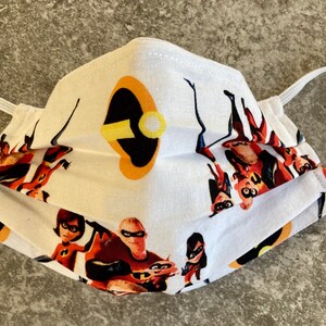 May include: White face mask with a colorful print of the Incredibles family. The mask has a white background with the Incredibles family in their superhero costumes. The mask also has a black and yellow symbol of Mr. Incredible's mask.