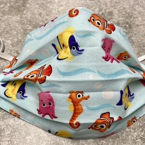 May include: A blue fabric face mask with a repeating pattern of cartoon sea creatures, including a seahorse, clownfish, octopus, and yellow fish. The mask has white elastic ear loops.