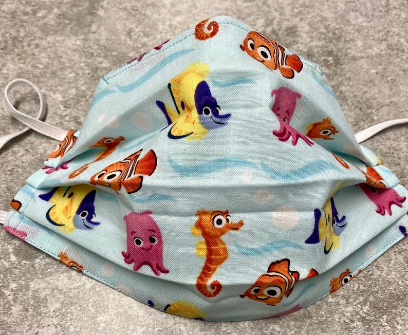 May include: A blue fabric face mask with a repeating pattern of cartoon sea creatures, including a seahorse, clownfish, octopus, and yellow fish. The mask has white elastic ear loops.