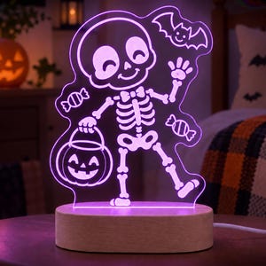 May include: A purple LED acrylic night light featuring a cartoon skeleton wearing a bowtie, holding a jack-o'-lantern bucket, and surrounded by two sweets and a flying bat. The light is mounted on a smooth, oval-shaped light-coloured wooden base.