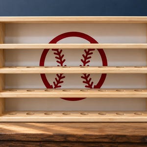 May include: A rectangular light-colored wooden display shelf featuring four horizontal tiers with circular cutouts for holding baseballs. The back panel is white and features a large, centered red graphic of a baseball with stitching.