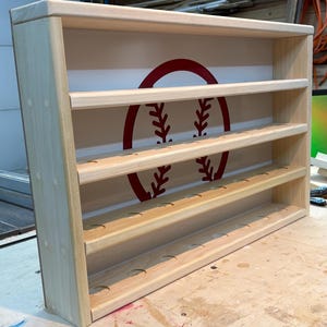 May include: A light-colored wooden display shelf featuring three horizontal tiers with circular indentations for holding items. The back panel is white and features a large, centered red graphic of a baseball with visible red stitching details.