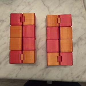 May include: Two orange and red infinity cubes are displayed side-by-side. Each cube is made of eight connected blocks. The cubes are on a white and gray marble surface. The cubes are a fun toy.