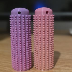 May include: Two 3D-printed, cylindrical objects with a textured surface of small, triangular spikes. One is lavender, the other is light pink. Each has a rounded top with a small hole. The objects are displayed on a wooden surface.