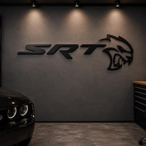 Può includere: Una grande insegna nera con il logo SRT Hellcat montata al centro di una parete grigio scuro. L'insegna presenta le lettere SRT seguite da un emblema stilizzato a testa di gatto. L'ambiente del garage include un'auto nera e vari attrezzi.