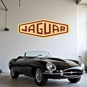 May include: A black Jaguar E-Type roadster parked in a garage with a vintage Jaguar logo on the wall behind it.