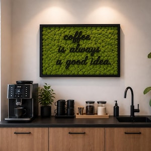 Könnte beinhalten: Ein rechteckiges Wandbild mit leuchtend grünem, konserviertem Moos in einem schwarzen Rahmen. Der Text "coffee is always a good idea." ist in schwarzen, dreidimensionalen Buchstaben mittig auf der Moosoberfläche angebracht.