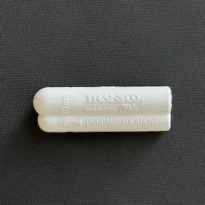 Puede incluir: Accesorio de tubería de plástico blanco con la etiqueta "TRANSCO®" con el texto "MADE IN CHINA C/W US" y "UP DRY/DAMP LOCATIONS". El accesorio de tubería tiene un diámetro de 10 mm.