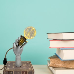 May include: A silver robotic hand holding a clear glass light bulb with warm white string lights inside. The light bulb is on a stack of books with a brown leather cover. The background is a light blue wall.
