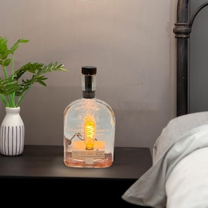 May include: A clear glass bottle lamp with a black cap and a coiled light bulb inside. The bottle is labeled "Woodford Reserve Kentucky Straight Bourbon Whiskey".