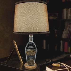 May include: A table lamp made from a clear glass bottle with a brown wooden base. The bottle is filled with small rocks and has the label "Angel's Envy" on it. The lamp has a beige fabric lampshade with a brown trim.