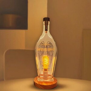 May include: A clear glass bottle lamp with a wooden base. The bottle has a design of angel wings etched on the front. The lamp is lit with a warm white light bulb.