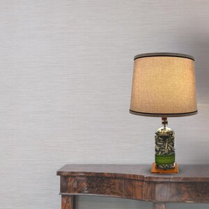 May include: A table lamp with a beige fabric lampshade and a clear glass bottle base filled with bullet casings. The bottle has a green label that reads "Bullets of War". The lamp is sitting on a dark brown wooden table.