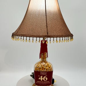 May include: A unique table lamp crafted from a Maker's Mark bottle, filled with corn kernels. The lamp features a brown lampshade with a decorative pattern and beaded trim. The bottle is mounted on a wooden base, with the text "46 FRENCH OAKED" visible.