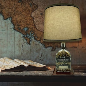 May include: A lamp made from a repurposed Woodford Reserve bourbon bottle with a beige fabric lampshade. The bottle is filled with small stones and the lamp is sitting on a wooden base.