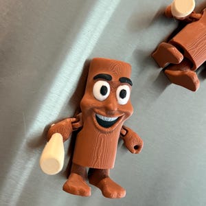 May include: A brown, cartoon-like figure with a smiling face, black eyebrows, and large white eyes. The figure is holding a beige object in its right hand. The figure has a wood grain texture and is standing on a metallic surface.