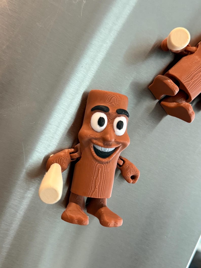 May include: A brown, cartoon-like figure with a smiling face, black eyebrows, and large white eyes. The figure is holding a beige object in its right hand. The figure has a wood grain texture and is standing on a metallic surface.