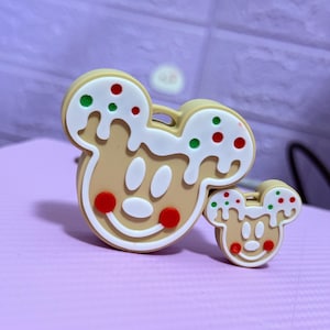 May include: Two Mickey Mouse-shaped gingerbread ornaments. The larger ornament is tan with white icing and red and green sprinkles. The smaller ornament has the same design. Both have a smiling face with red circles for cheeks.