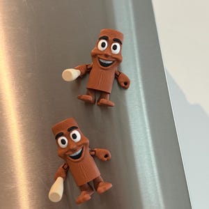 May include: Two brown, cartoon-like figures with smiling faces and white cylindrical objects in their hands. The figures have black eyes and are posed as if they are waving. They are set against a brushed metal background.