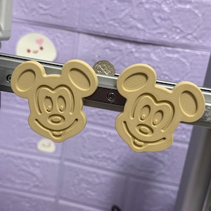 Może przedstawiać: Dwa wykrawacze do ciastek w kształcie Myszki Miki w kolorze beżowym. Wykrawacze wykonane są z wytrzymałego materiału i przedstawiają kultową twarz myszy z dużymi uszami, uśmiechniętymi ustami i okrągłymi oczami. Tło to jasnofioletowa ceglana ściana.