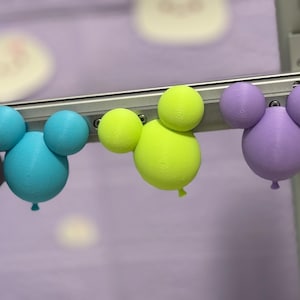 Op de afbeelding: Drie kleurrijke ballonvormige decoraties in blauw, geelgroen en paars. Elk heeft twee kleinere bollen bovenop, die lijken op een stripfiguur. De decoraties zijn bevestigd aan een metalen staaf.