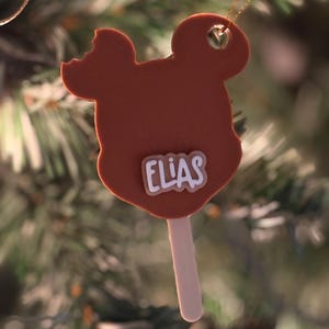 May include: A brown, Mickey Mouse-shaped Christmas tree ornament with a bite taken out of the ear. The name "ELIAS" is written in white letters on the ornament. A wooden stick is attached to the bottom. A gold chain is attached to the top for hanging.