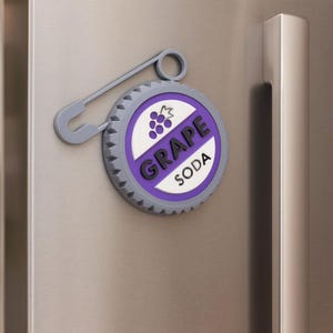 May include: A gray refrigerator magnet shaped like a grape soda bottle cap. The magnet has a purple and white design with the words "GRAPE SODA" and a grape cluster graphic. A gray safety pin is attached to the magnet.