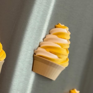 Może przedstawiać: Zbliżenie magnesu w kształcie rożka z lodami. Lody mają wirujący wzór w kolorach żółtym i białym. Stożek ma jasnobrązowy kolor.