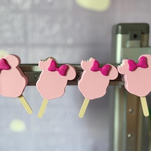Könnte beinhalten: Vier rosa Minnie-Maus-förmige Leckereien mit leuchtend rosa Schleifen und Holzstäbchen. Einige Leckereien weisen Bissspuren auf. Die Leckereien werden auf einer Metallstange vor einem verschwommenen Hintergrund präsentiert.