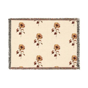 May include: Cream-coloured woven throw blanket with a repeating pattern of sunflowers in yellow, brown, and green. The blanket has a fringed edge in neutral tones. The sunflowers are depicted with detailed petals and stems.