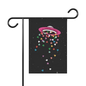 May include: A black garden flag with a pink UFO design, releasing colourful heart shapes against a starry backdrop. The flag is mounted on a black metal stand with a curved top.