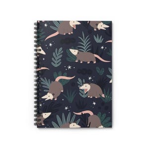 Peut inclure: Un carnet à spirales bleu foncé avec un motif répété d'opossums et de feuillage. Les opossums sont illustrés dans un style cartoon avec des nez roses et des dents blanches. Le feuillage est un mélange de vert et de bleu.