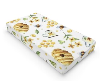 May include: A white fitted cot sheet with a repeating pattern of honeycombs, bees, flowers, and honey pots. The pattern is in shades of yellow, brown, and green.