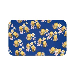 May include: A rectangular, blue bath mat with a repeating pattern of cartoon bees, white daisies, and pink flowers. The bees are yellow and black, with smiling faces. The flowers are scattered across the surface.