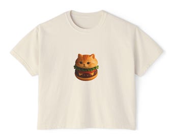 May include: Cream-colored t-shirt featuring a whimsical graphic of a cat's head integrated into a hamburger design. The cat-burger has a bun, lettuce, tomato, and a patty. The shirt has a crew neck and short sleeves, suitable for casual wear.