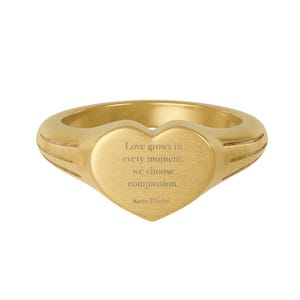 Könnte beinhalten: Ein goldfarbener Ring mit herzförmiger Oberfläche. Der Ring trägt die Inschrift "Love grows in every moment, we choose compassion. Savor Hozho" auf dem Herzen. Der Ring ist ein Schmuckstück.