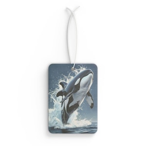 May include: Rectangular air freshener featuring a leaping orca whale illustration. The orca is black and white, emerging from the ocean with a splash. The background is a gradient of blue hues, and a white string is attached.