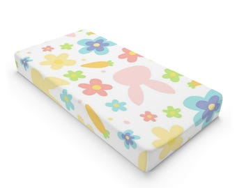 May include: A white fitted cot sheet with a pastel floral and bunny pattern. The sheet features pink bunnies, yellow, blue, and green flowers, and orange carrots.