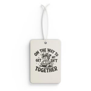 May include: Rectangular air freshener with a white background and a string for hanging. The design features a raccoon wearing a cowboy hat and sunglasses, riding a turtle. Text reads "ON THE WAY TO GET MY SH*T TOGETHER."
